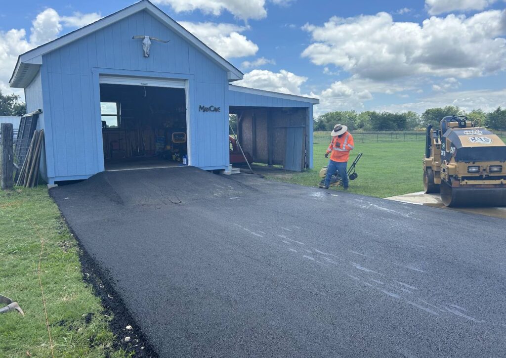 Paving worker using a plate compactor alongside a CAT roller on a newly installed asphalt driveway ramp in Heath TX