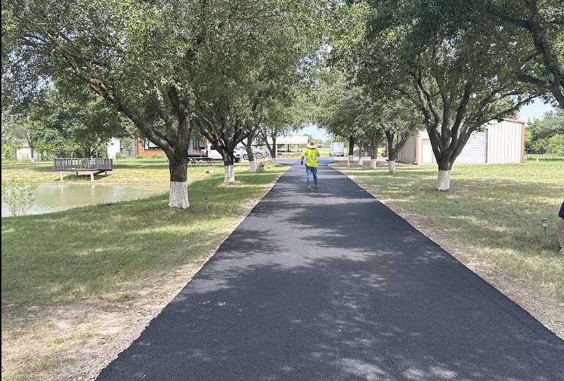 Newly paved blacktop residential driveway running under shade trees alongside a pond and barn in Rowlett TX.