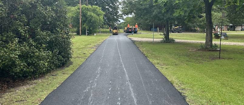 Newly paved residential asphalt driveway extending through a grassy tree-lined yard with paving equipment working in Fate TX.