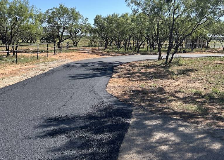 Newly paved blacktop driveway curving through a rural property surrounded by mesquite trees and ranch fencing in Forney TX.