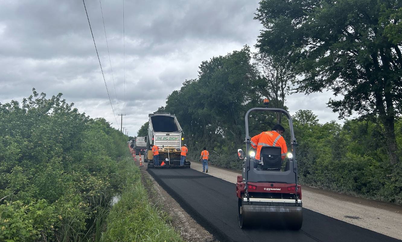 Dynapac roller compacting fresh asphalt behind a paving train crew on a rural road in Talty TX by Gentry's Paving Group.